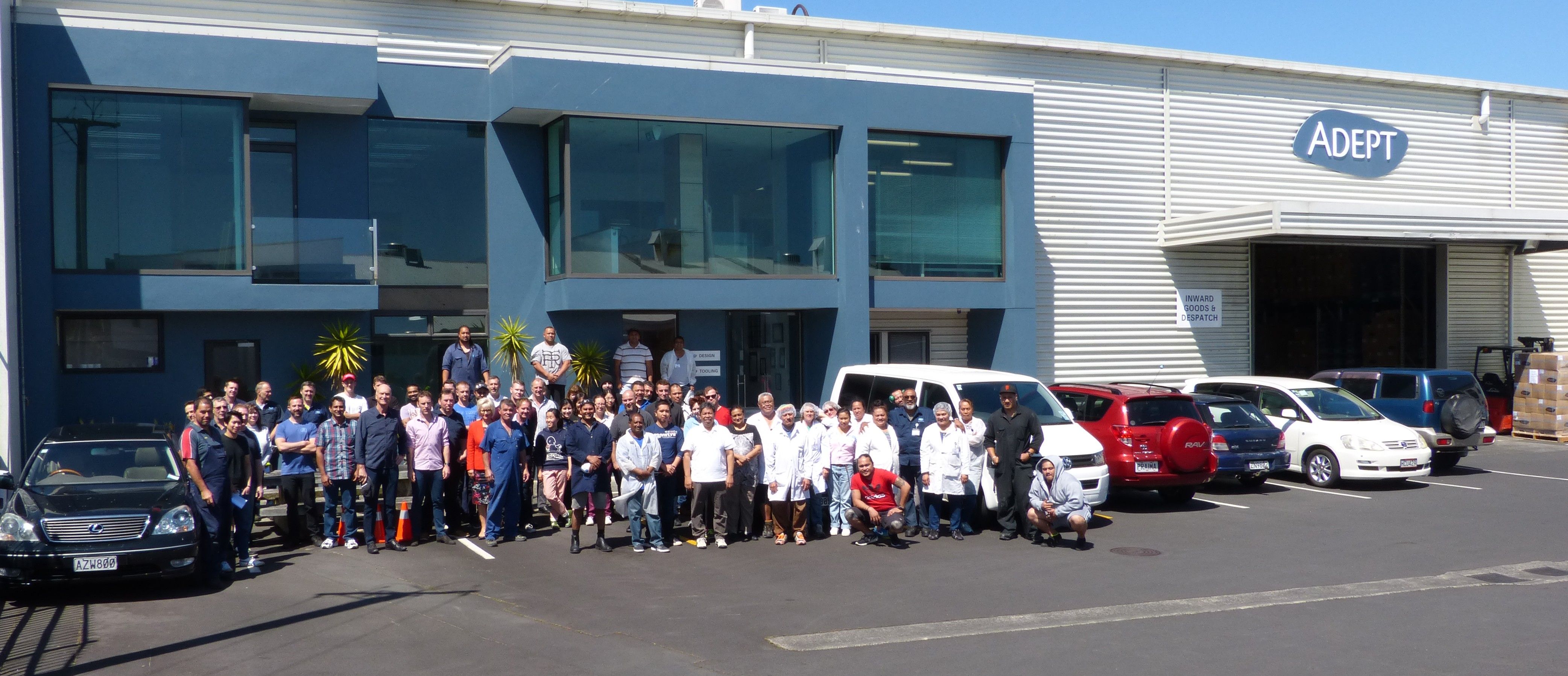 A large group of staff stands in front of the Adept company building and warehouse with several cars parked.