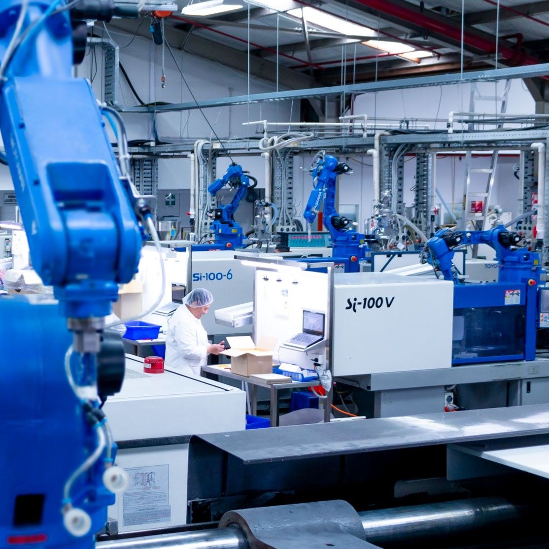 A manufacturing facility with blue robotic arms and automated machines, where a technician in a lab coat inspects items.