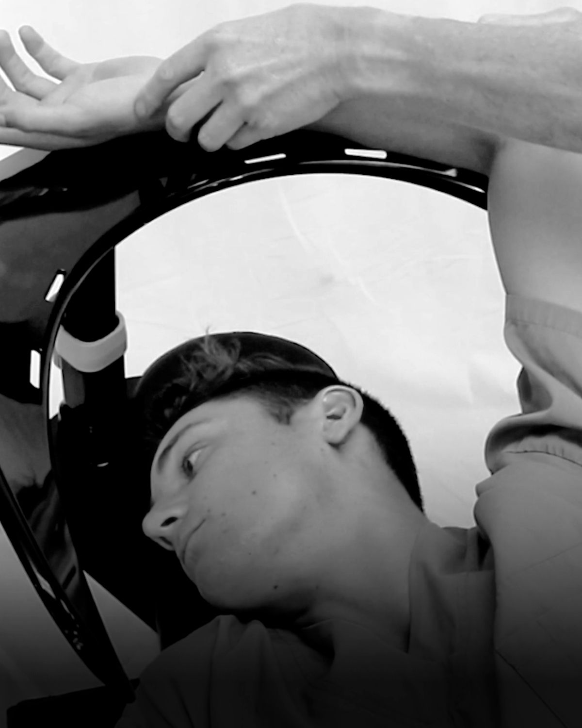 Black and white image of a young man lying down, observing a hand examining a wrist supported by a curved medical device.